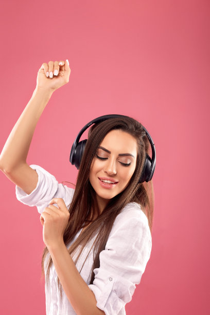 在粉色背景下戴无线耳机唱歌的漂亮棕发女士图片下载