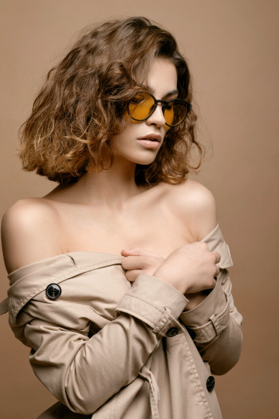 美丽的时尚模特与干净的皮肤和卷发在米色斗篷拉伸米色背景，模特在时尚眼镜，裸露的肩膀图片下载