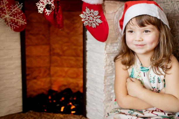 可爱的微笑小女孩戴着圣诞帽在美丽的客厅与传统的壁炉装饰圣诞节。图片下载