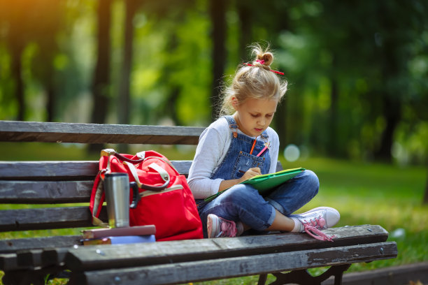 在阳光明媚的公园里，一个漂亮的小女孩坐在长凳上用彩色铅笔画画图片下载