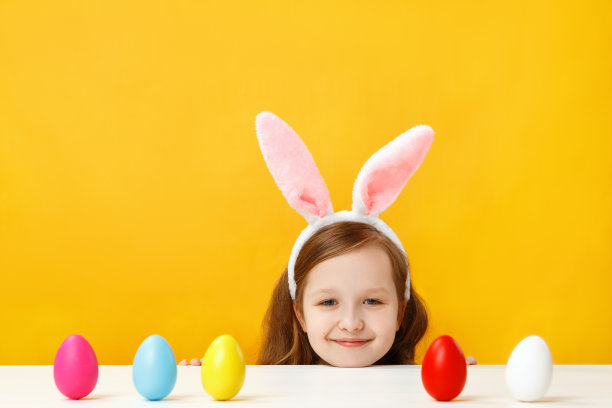 复活节的概念。快乐的婴儿在兔子的耳朵上黄色的背景。一个小女孩从桌子上往外看，寻找彩蛋图片下载