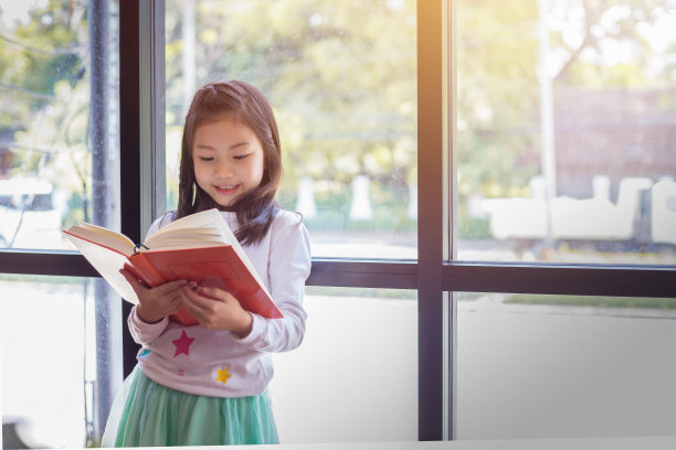 一个亚洲女孩站在窗边读一本红书。图片下载