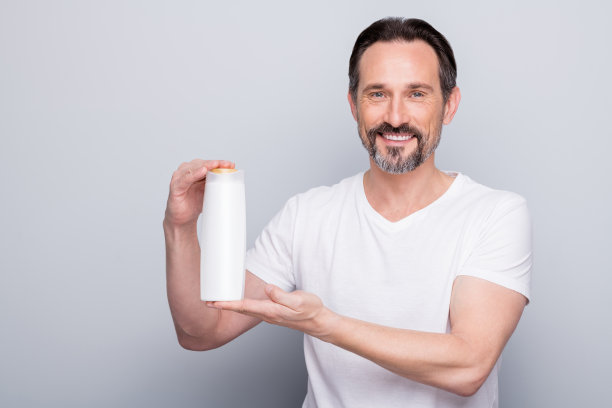 照片英俊的老男人男人拿着洗发水护发素瓶子两在一个惊人的效果，广告买家穿白色t恤孤立的灰色背景图片下载