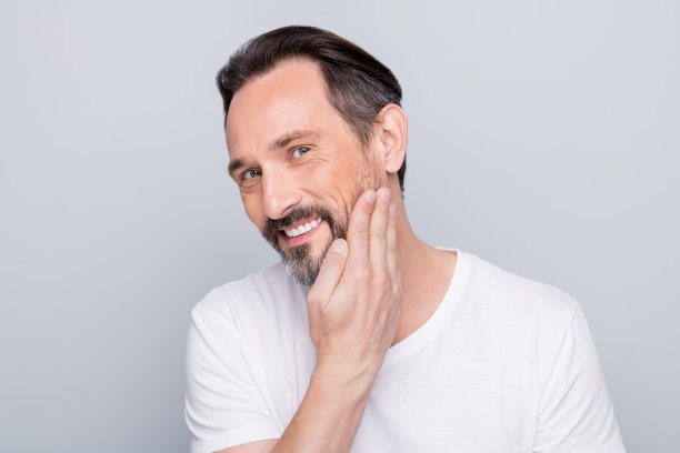 英俊积极成熟的男人的照片看着镜子，牙齿微笑的手检查丝绸般的皮肤后，使用剃须凝胶穿白色t恤孤立的灰色背景图片下载