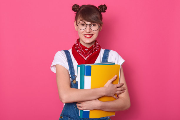 快乐好看的学生特写，拿着彩色纸质文件夹，戴着头巾和T恤，穿着背带裤，看着镜头微笑，发型有趣，在玫瑰色背景下摆姿势图片下载