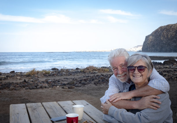 一对快乐的老年夫妇在海滩上拥抱。坐在一张木桌旁，喝着两杯咖啡。两个真实的人图片下载