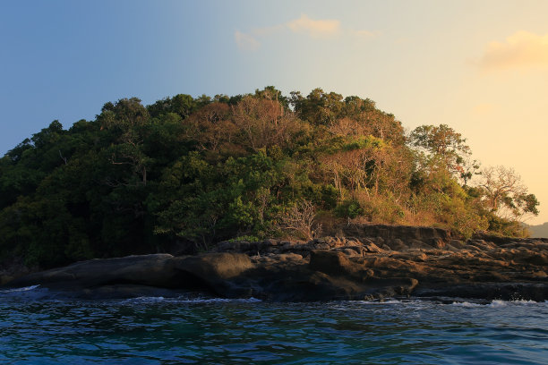 海上五颜六色的日出。橘黄色的晨光投射在海洋中央的岛屿森林上。森林里的树木丛林覆盖着岩岸，海浪轻柔地滚向石头。图片下载