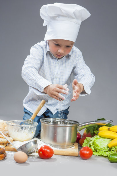美食概念：专心致志的白人男孩正在用面粉工作，用筛子将其筛到平底锅上。戴着厨师帽，周围环绕着水果和蔬菜，以灰色为背景的竖版照片图片下载