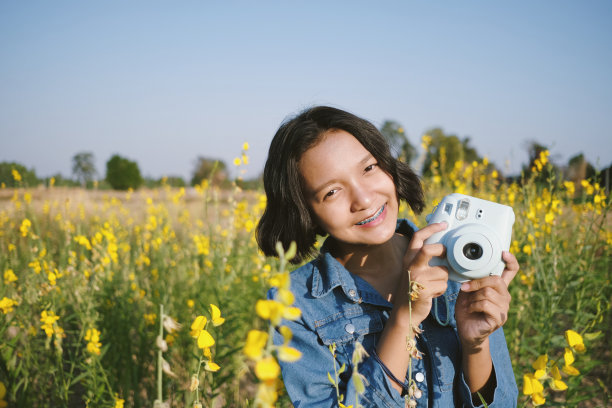 可爱的快乐亚洲年轻女孩使用相机穿牛仔裤在美丽的黄花花园。图片下载