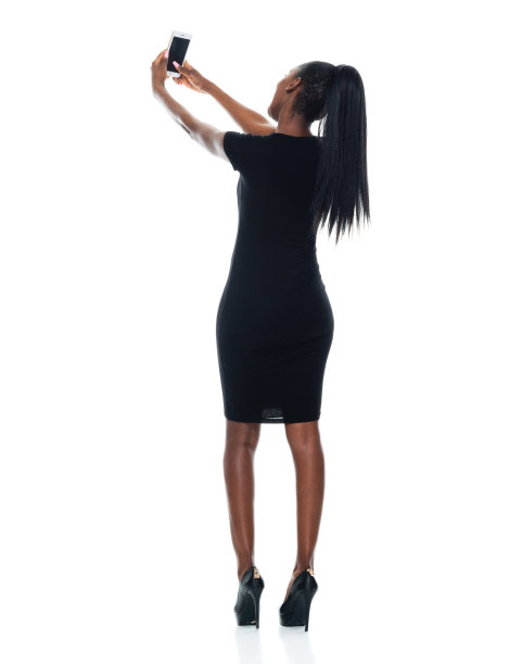 非裔美国女性穿着裙子，使用手机，站在白色背景前拍照图片下载