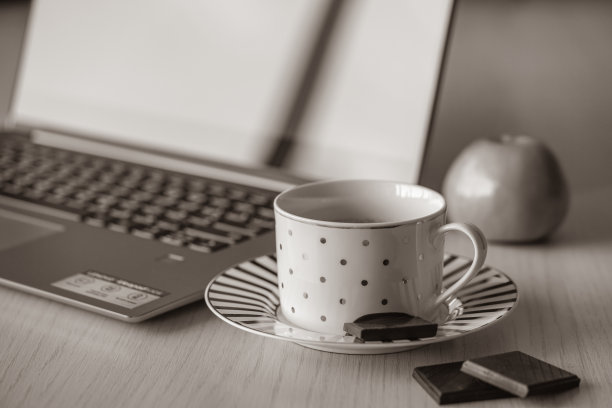 木桌上放着一杯咖啡，背景是笔记本电脑和苹果。在办公室的休息时间放上一块黑巧克力。黑白图像。商业静物图片下载