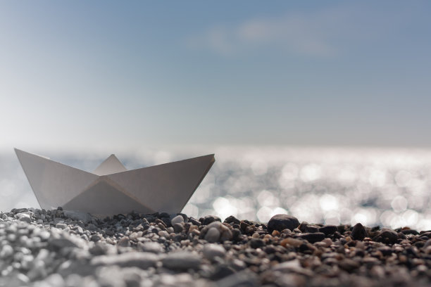 在一个阳光明媚的日子里，一艘纸船在一个鹅卵石海滩上。图片下载