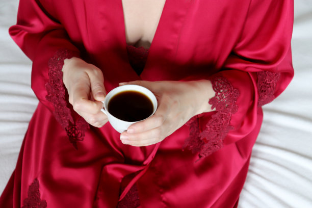 一杯咖啡在女人的手中，性感的女人在丝绸红色睡衣坐在床上图片下载