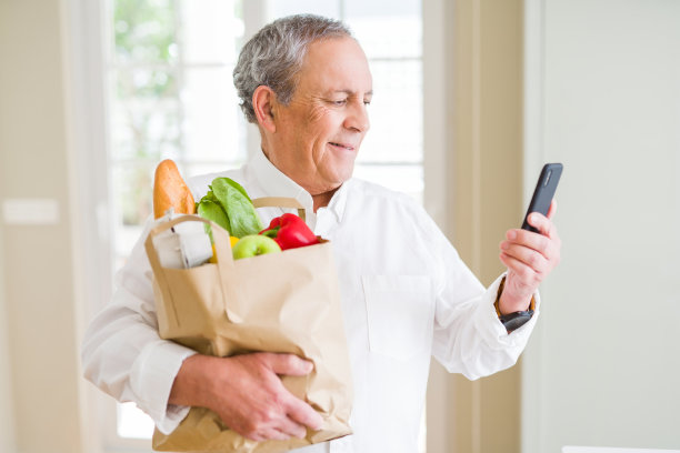 英俊的老男人拿着装满新鲜食品的纸袋，看着智能手机微笑图片下载