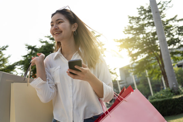 美丽的女孩长头发她是快乐的购物在街上的商场，她拿着几袋纸和手机，光散拍复制空间的照片。图片下载