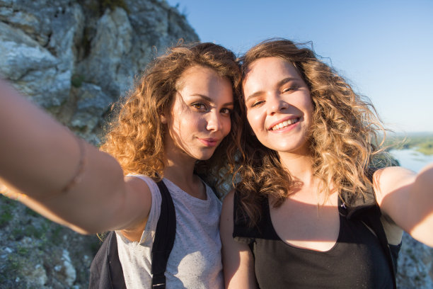 一个没有国界的世界。两个女孩在山里的惊人旅程。两个迷人的年轻女孩一起旅行，在阳光下对着天空和山中的风景用手机自拍图片下载