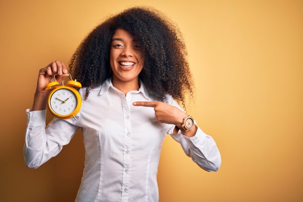 年轻的非洲裔美国妇女与非洲式的头发拿着经典的闹钟在黄色的背景非常幸福的指与手和手指图片下载
