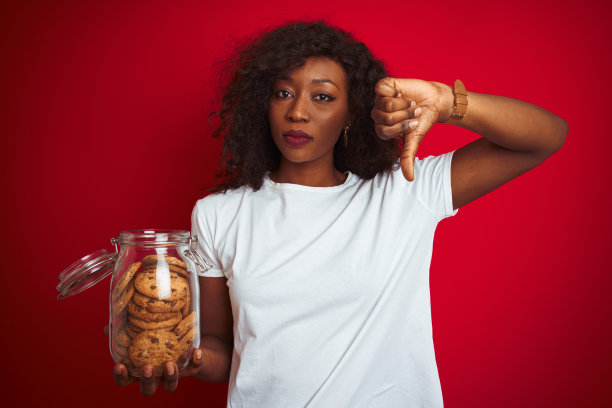 年轻的非洲裔美国妇女拿着一罐饼干在孤立的红色背景下愤怒的脸，消极的标志表示不喜欢，拇指下来，拒绝的概念图片下载
