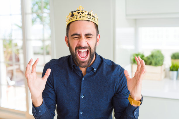 英俊的西班牙裔男子头上戴着金色皇冠，国王高举双臂，闭着眼睛兴奋地尖叫，庆祝疯狂的成功。赢家的概念图片下载