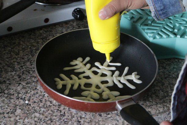 制作圣诞薄饼的图片，用挤压酱汁瓶为圣诞节儿童早餐制作的滚轴状雪花煎饼，用不粘锅在灶台上烹饪的雪花煎饼，概念图片。图片下载