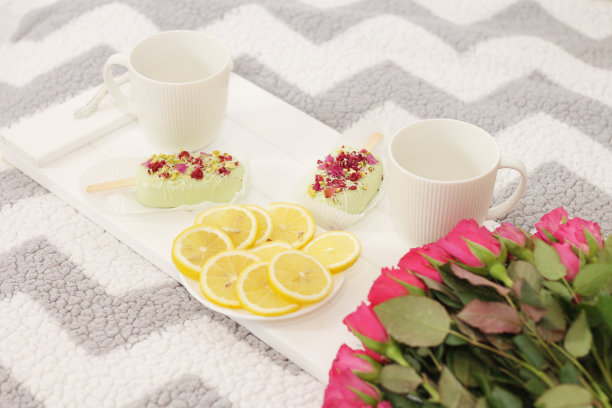 一个白色木制托盘，上面有两个杯子，两个冰棒蛋糕，一个盘子，上面有切好的柠檬和一束粉红色的玫瑰。图片下载