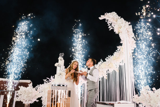新娘和新郎正在切婚礼蛋糕。手用鲜花切蛋糕。新婚夫妇在码头拱门附近。夜晚的仪式有灯光和烟花。火表演。图片下载