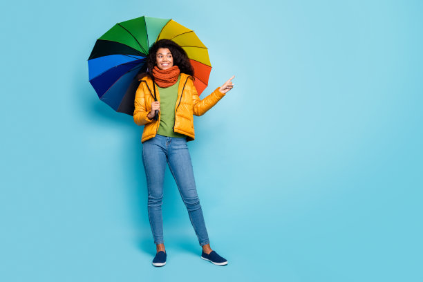 美丽的黑皮肤女士的全尺寸照片拿着彩色伞指示手指空的新奇产品穿大衣牛仔裤毛衣围巾孤立的蓝色背景图片下载