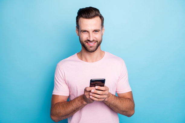 有吸引力的家伙握着电话的手的照片博主写新的评论现代科技的人穿休闲的粉红色t恤孤立的蓝色背景图片下载