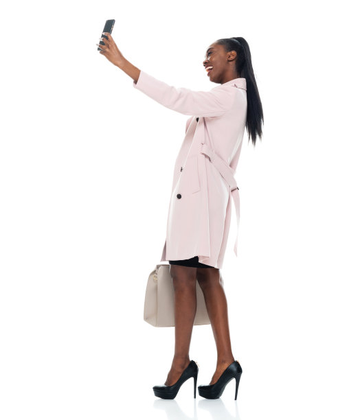 非洲裔女性穿着风衣站在白色背景前，拿着钱包和使用手机图片下载