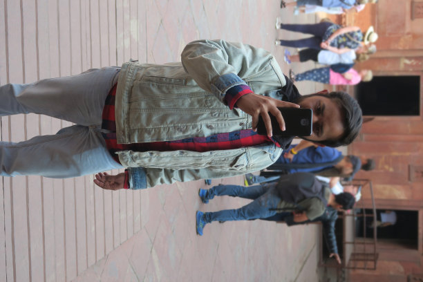 年轻英俊的印度男子在20岁出头用手机相机自拍，举着手机，看着手机屏幕自拍自拍在历史悠久的莫卧儿阿格拉堡与游客在背景阿格拉，印度男子的头发梳到后面图片下载