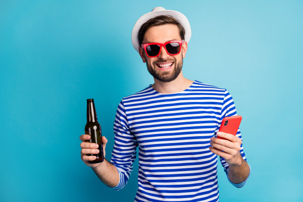 时髦的好旅行者的照片男人浏览电话喝啤酒瓶所有包括异国情调的度假胜地戴太阳眼镜条纹水手衬衫帽孤立的蓝色背景图片下载