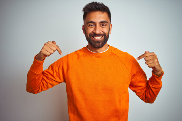 自信微笑的年轻印度男子，身穿橙色毛衣，指向自己表达自豪和喜悦图片下载
