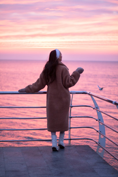 身穿皮草大衣的年轻女孩在海滨长廊欣赏海上日出，手捧热饮图片下载