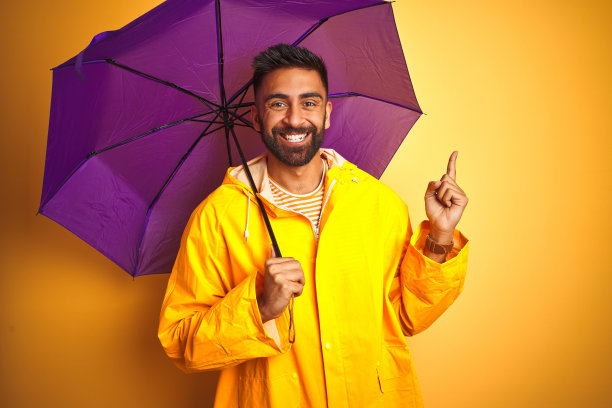 穿着雨衣，拿着紫色雨伞的年轻印度男子，在黄色背景前高兴地用手和手指指向一侧图片下载