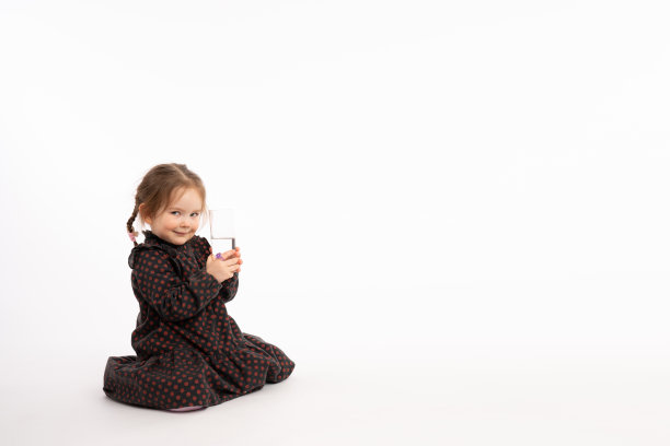 自信的红发儿童身穿红色豌豆连衣裙坐在地板上，手拿一杯水看着镜头，白色背景图片下载