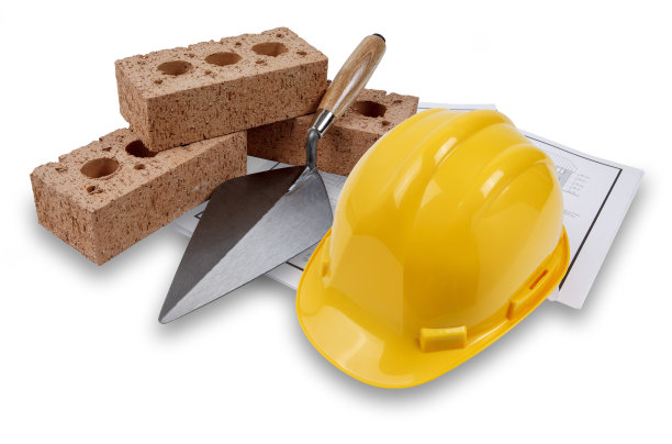 一套房子的建筑用砖和泥铲，戴着一顶黄色的安全帽，在一套建筑平面图上，孤立在白色的阴影下图片下载