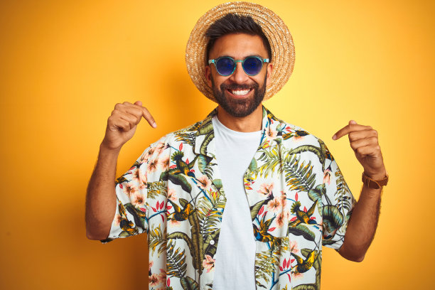 印度男子在黄色背景度假，戴着花衬衫帽子和太阳镜，面带微笑，自信地指着自己图片下载