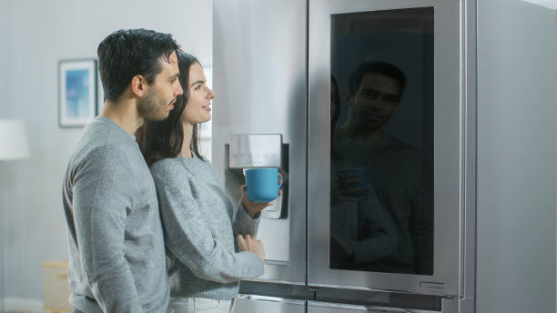 一对美丽的年轻夫妇在厨房里喝早上的咖啡。他们在家里的智能冰箱上查看天气预报和待办事项清单。公寓明亮舒适。图片下载