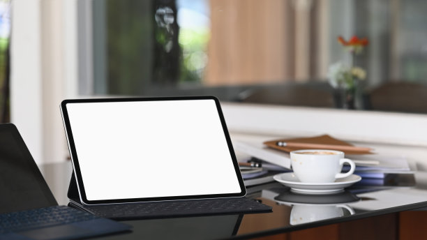 黑屏带键盘壳的平板电脑放在现代的工作台上，以咖啡杯和模糊的办公室为背景。图片下载