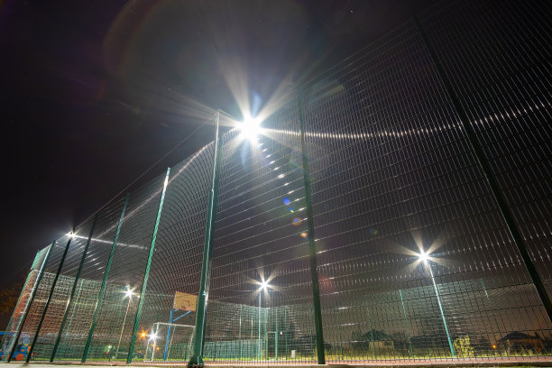 户外迷你足球场和篮球场，球门和篮筐环绕着高防护网，在夜晚被聚光灯照亮。图片下载