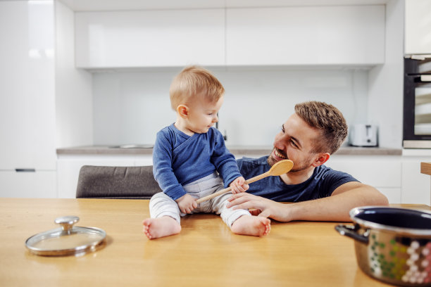 可爱的小男孩坐在厨房柜台上拿着搅拌勺，父亲蹲在他旁边，他们正在为家人准备饭菜图片下载