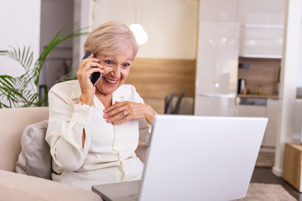 快乐的高级女人与笔记本电脑，坐在沙发上与某人在电话上交谈。使用新技术更容易也更快图片下载