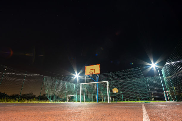 户外迷你足球场和篮球场，球门和篮筐环绕着高防护网，在夜晚被聚光灯照亮。图片下载