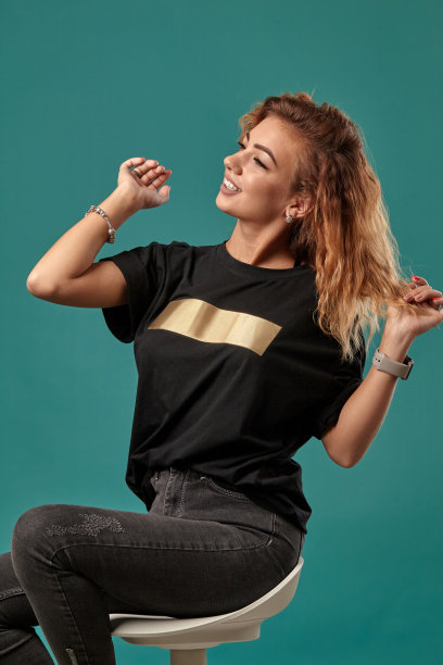 一头卷发的姜黄色女孩，穿着一件带有金色条纹的黑色t恤，正坐在椅子上摆造型，背景是工作室的蓝色。特写镜头图片下载