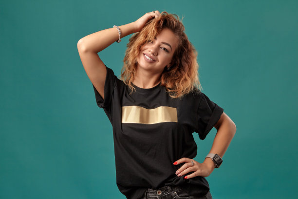 一个姜黄色的女孩，一头卷发，穿着黑色的t恤和金色的条纹是站在一个蓝色的工作室背景。特写镜头图片下载