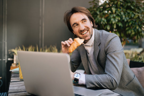 快乐的商人吃牛角面包，同时放松在户外咖啡馆。图片下载