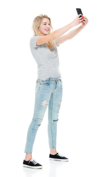 z一代的年轻女性穿着牛仔裤站在白色背景前用手机拍照图片下载