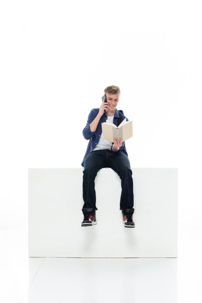 男大学生穿着牛仔裤在白色背景前休息，拿着书和使用手机图片下载