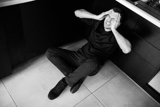 黑白照片：身穿黑衣的年轻男人独自坐在厨房里尖叫，双手抱头试图冷静，一片狼藉，充满痛苦和绝望。图片下载
