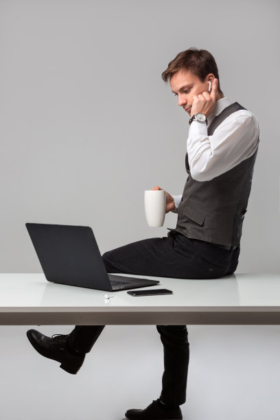 男人坐在放有笔记本电脑和智能手机的白色桌子旁喝咖啡图片下载